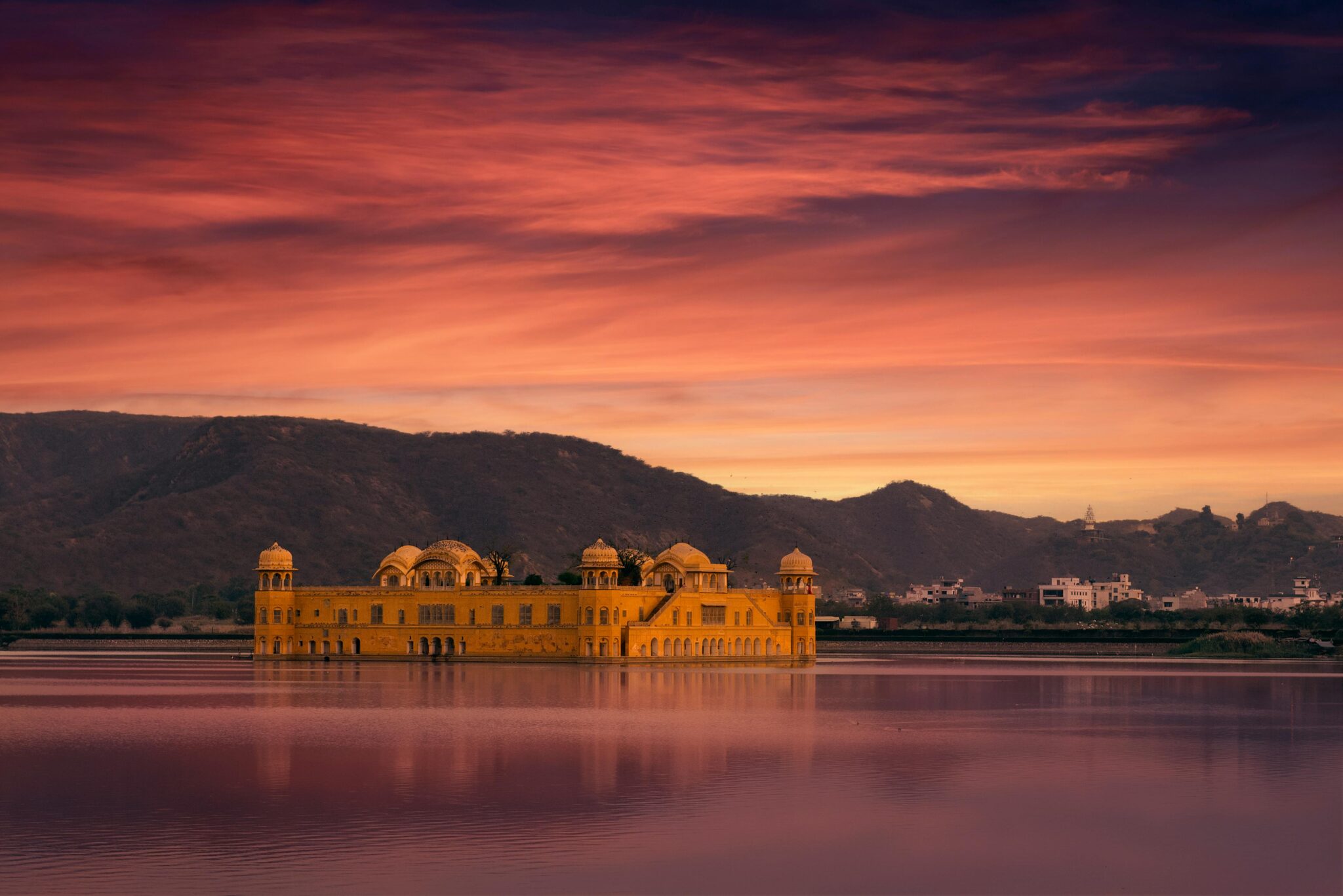 Stunning view of Jal Mahal palace reflecting in a serene lake under a dramatic sunset sky in Jaipur, India.